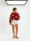 Dark red knit sweater with large white daisies, worn by a model with white ruffled shorts and a straw bag.