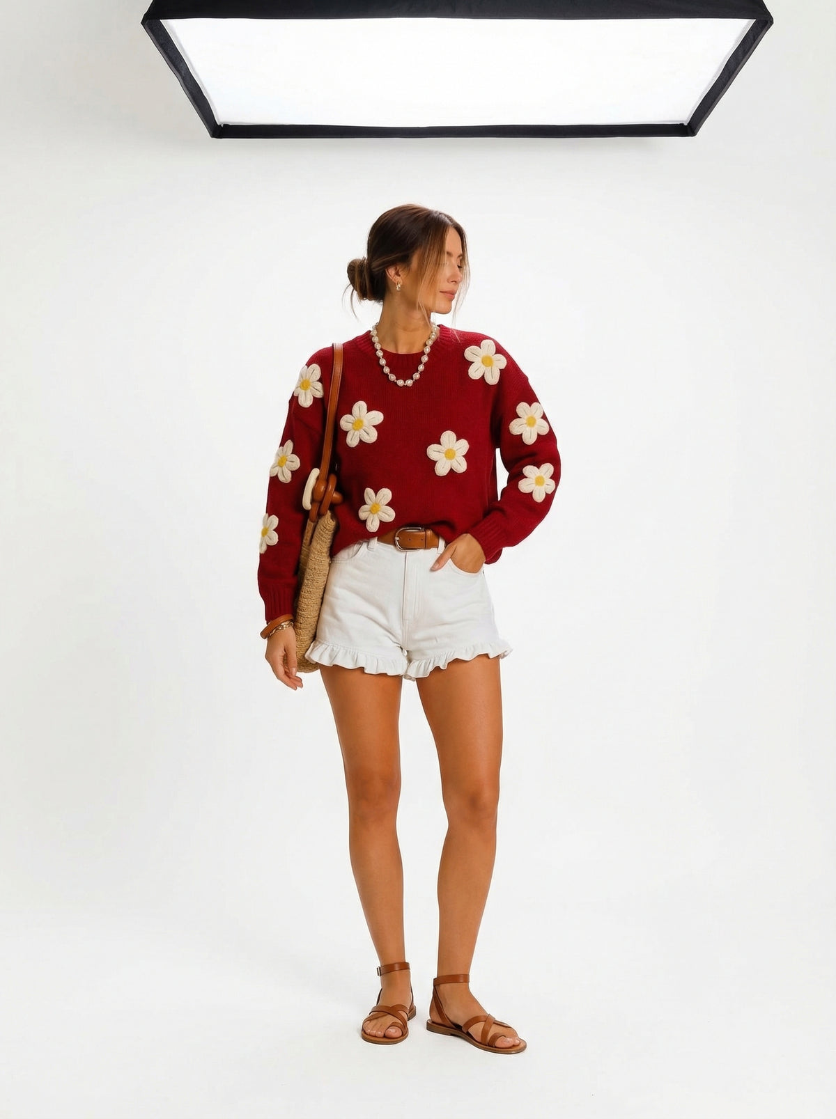 Dark red knit sweater with large white daisies, worn by a model with white ruffled shorts and a straw bag.