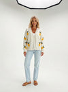 Woman wearing a cream V-neck knit cardigan with yellow lemons and green leaves, light blue jeans, and brown sandals.