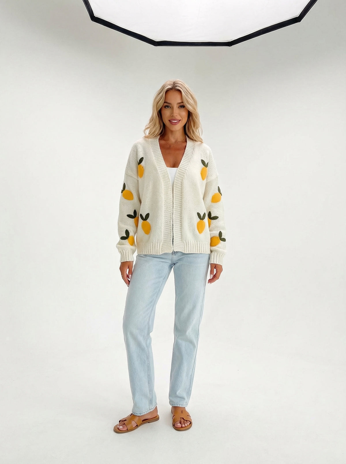 Woman wearing a cream V-neck knit cardigan with yellow lemons and green leaves, light blue jeans, and brown sandals.