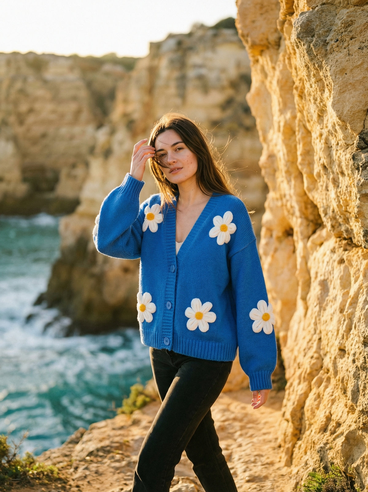 Woman wearing a bright blue knit V-neck cardigan with large white daisy appliques and front buttons, outdoors.