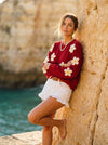 Woman wearing a red knit sweater with large white daisies and white ruffled shorts, leaning against a rock.
