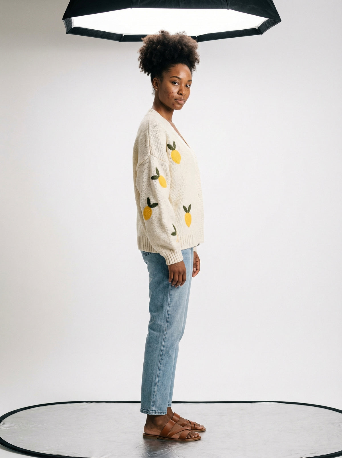 Woman standing in profile wearing a cream knit cardigan with yellow lemons, light blue jeans, and brown sandals.
